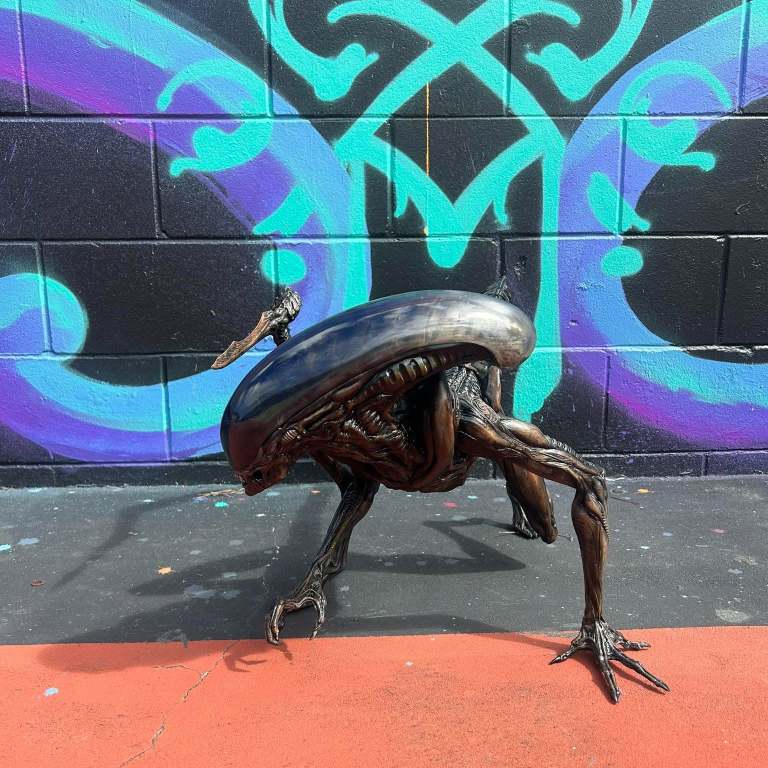 Bronze-finish 3D printed crawling xenomorph in front of a graffiti wall